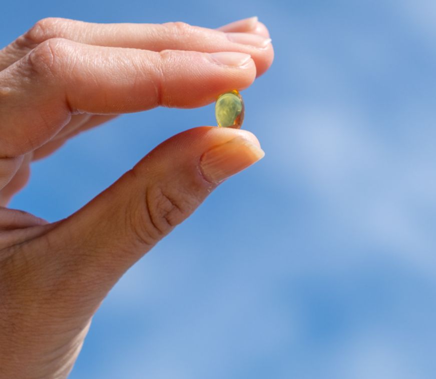 Una mano tiene un singolo Aavalabs Vitamina D3+K2 Softgel davanti a un cielo blu.