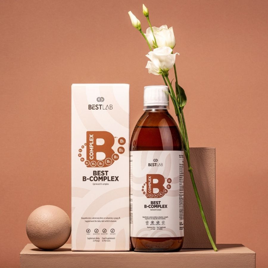 Bottiglia di vetro marrone e scatola "BEST LAB B-COMPLEX" su piedistallo in legno. Fiori bianchi e sfera decorativi.