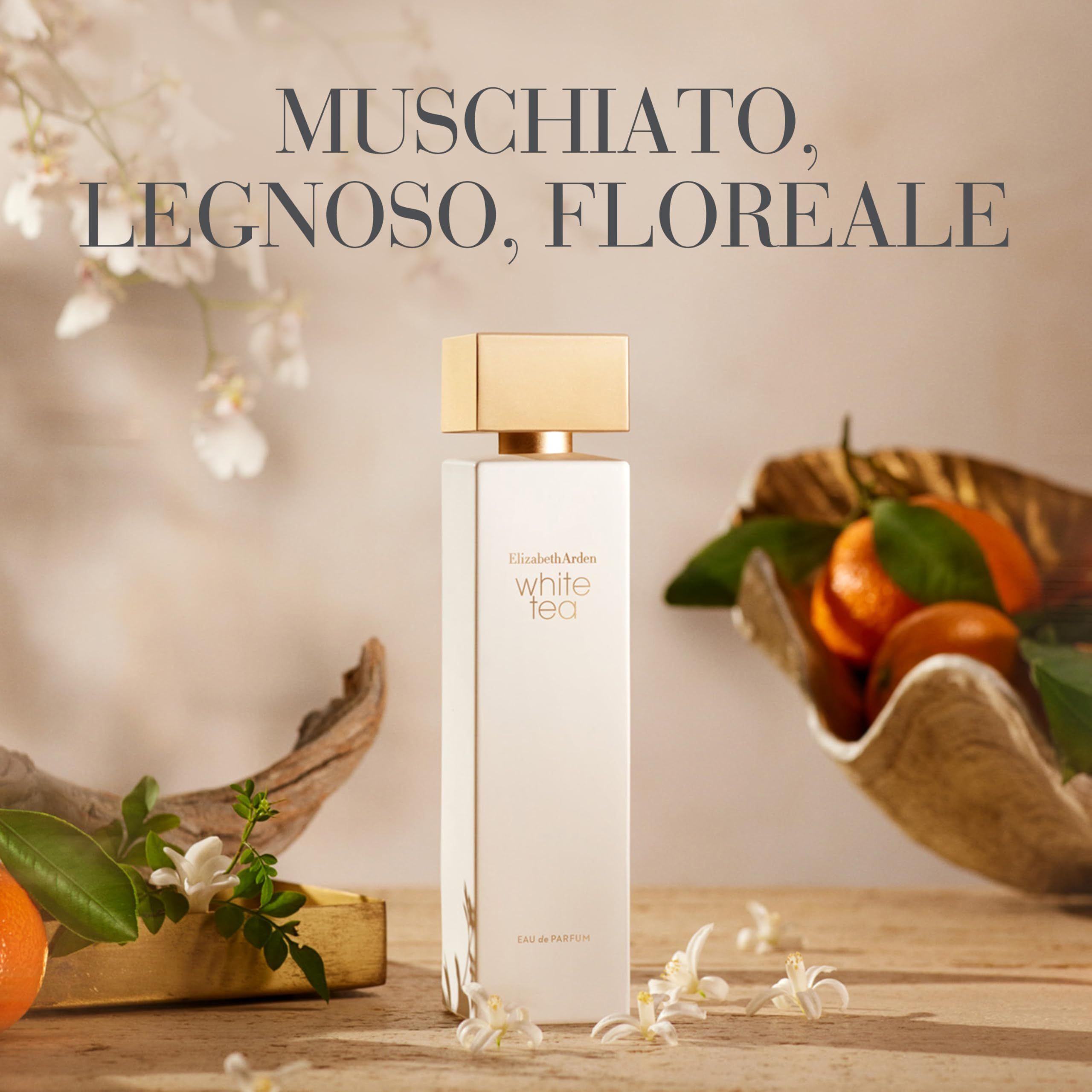 Flacone di profumo bianco con tappo dorato. Scritta: White Tea. Sfondo: legno, fiori, arance.