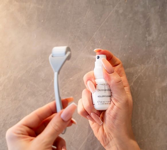 Mani che tengono Dermaroller e flacone spray. Flacone con scritta Roller Cleaner. Rullo grigio, flacone bianco.