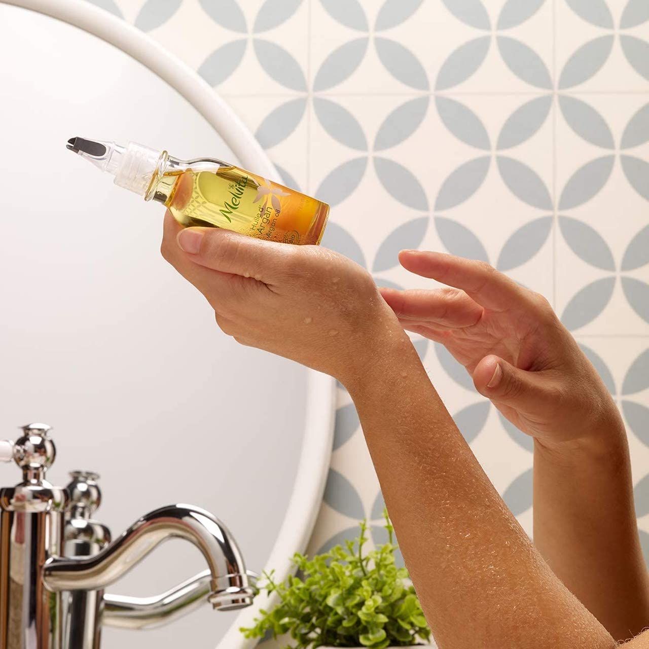 Mani che tengono un flacone di olio sopra un lavandino. Specchio sullo sfondo. Prodotto: Melvita Olio d'Argan Biologico.