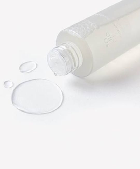 Liquido trasparente che esce da una bottiglia. Gocce e pozza su sfondo bianco. Bottiglia con scritta SOROCI.