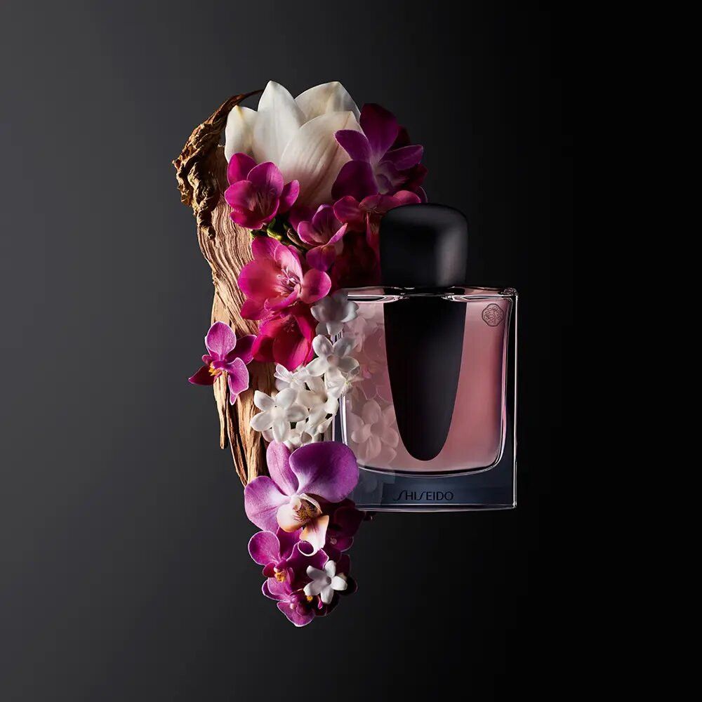 Flacone di profumo accanto a composizione floreale. Tappo nero, liquido rosa, logo Shiseido.