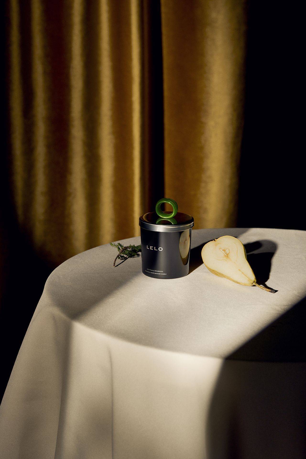 Barattolo nero con manico verde e pera su tavolo. Scritta LELO. Testo: Snow Pear & Cedarwood, FUCKING TOUCH MASSAGE CANDLE.