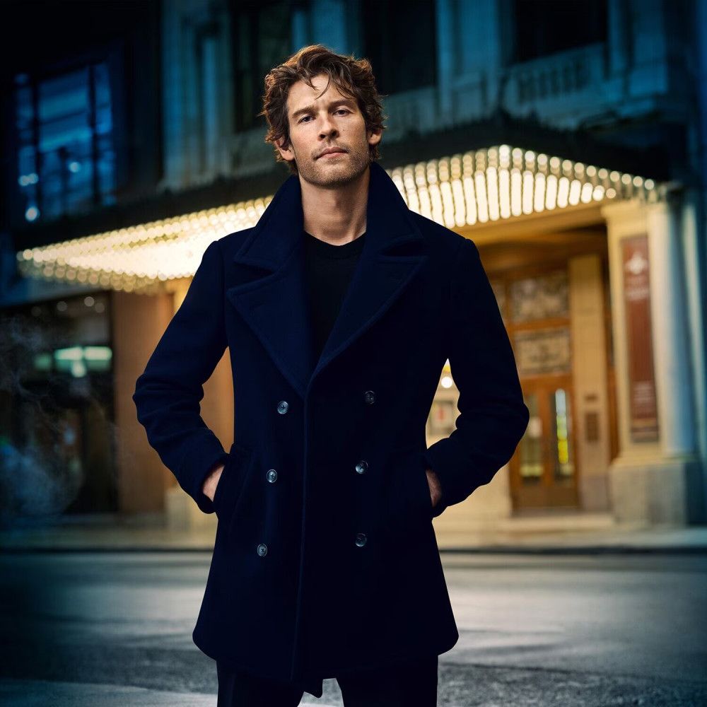 Uomo in cappotto blu scuro davanti a un edificio. Mani in tasca.