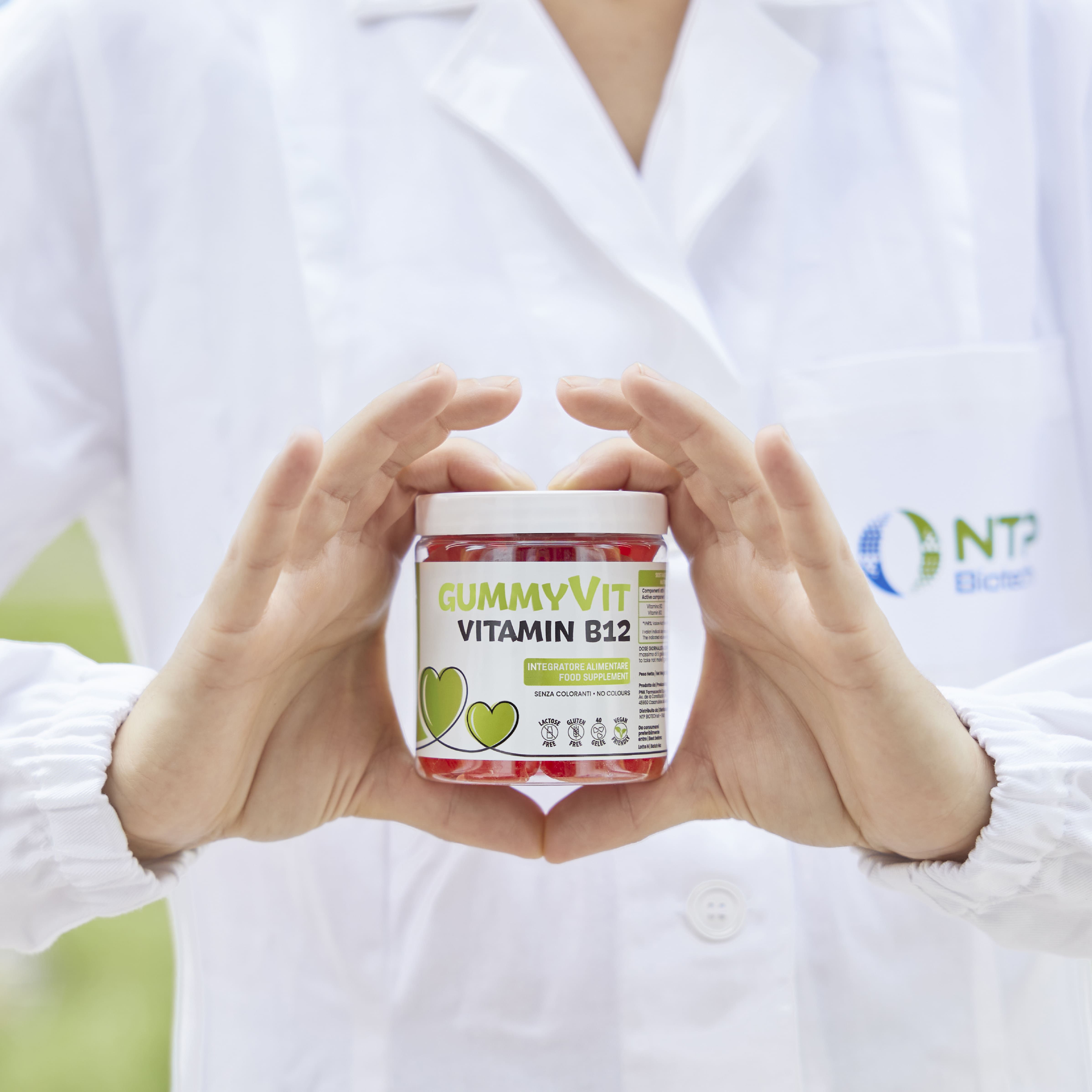 Persona che tiene un barattolo di Gummyvit B12. Scritta: Vitamina B12. Barattolo con caramelle gommose rosse. Logo NTP Biotech.