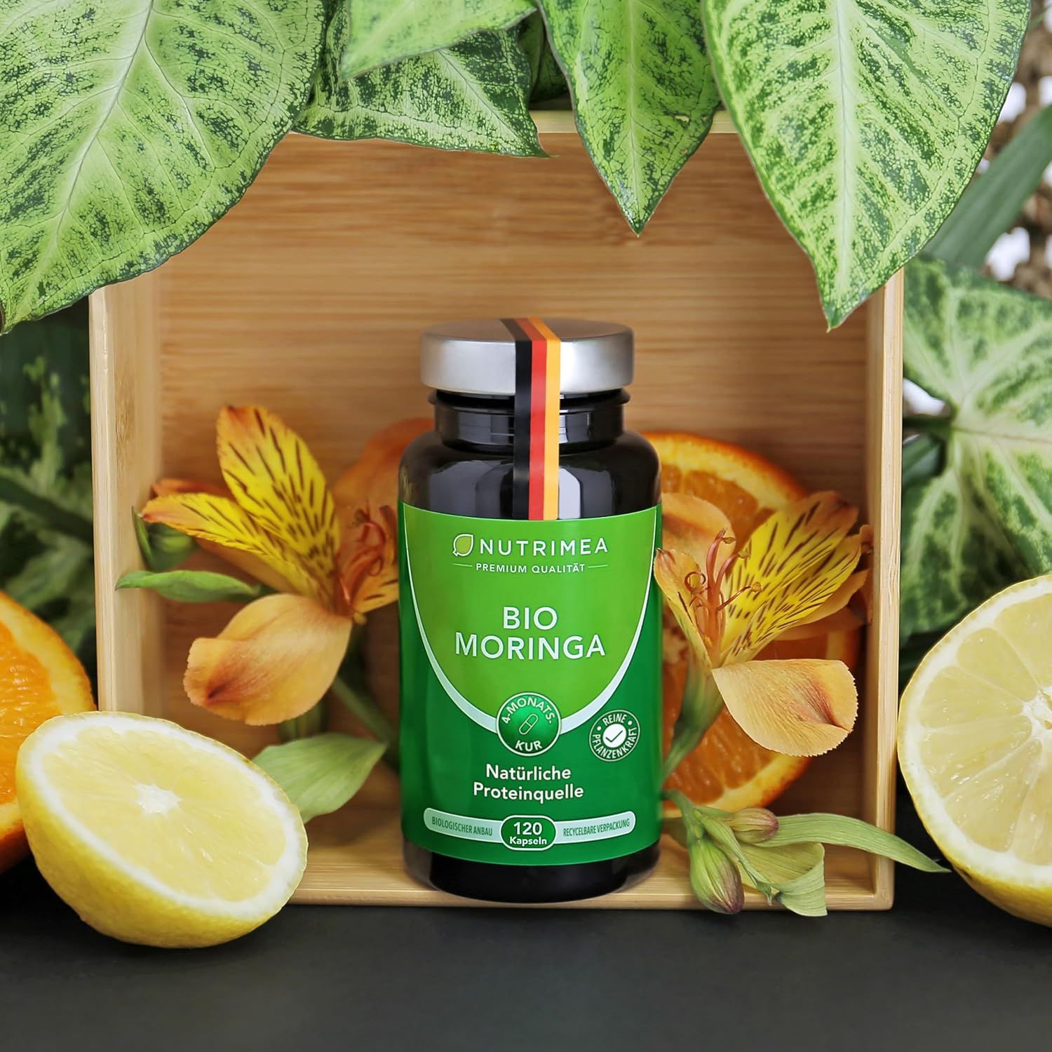 Bottiglia di capsule Nutrimea Bio Moringa, circondata da agrumi e foglie. Coperchio nero con bandiera tedesca.
