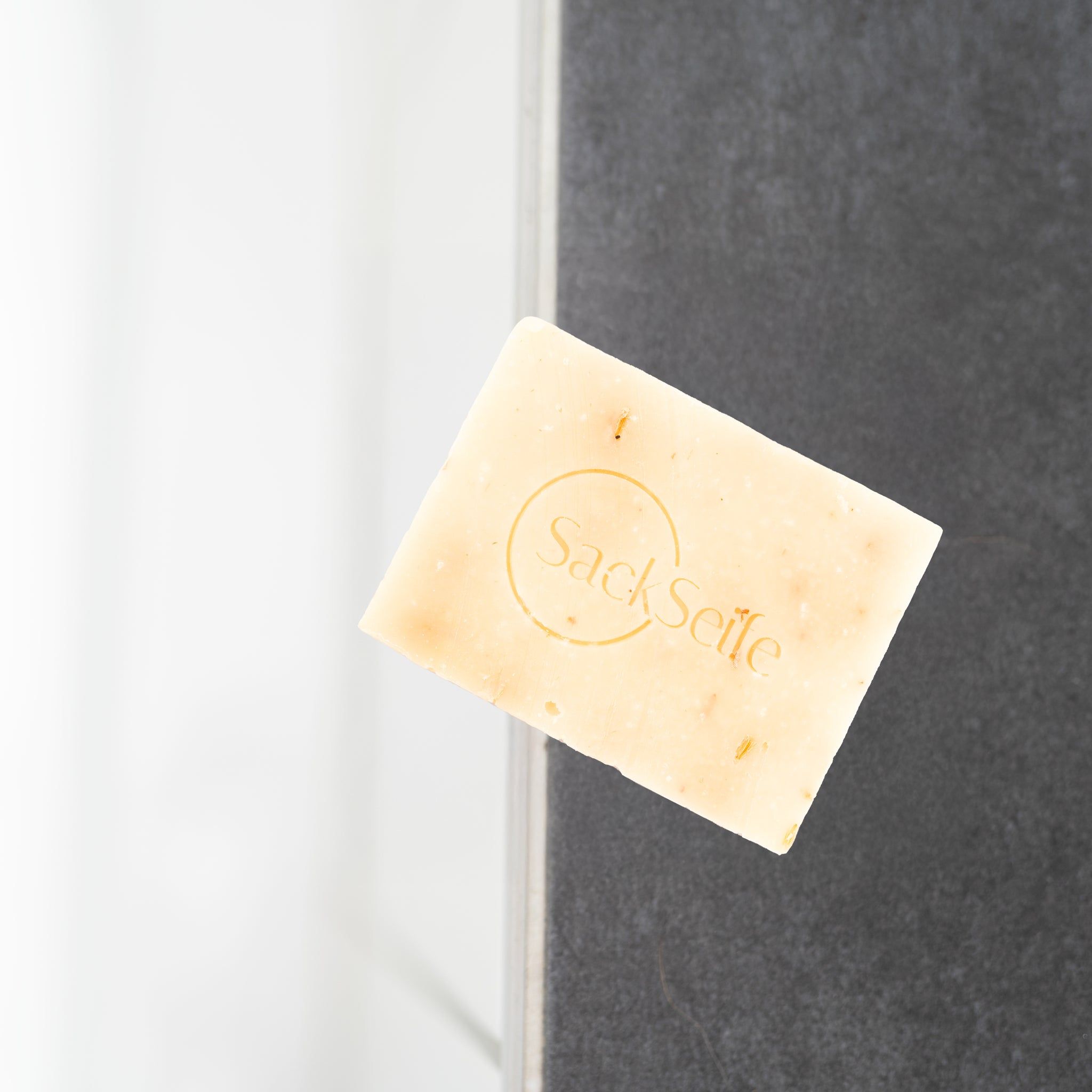 Sapone rettangolare con logo SackSeife. Colore grigio. Appoggiato a una parete grigia.