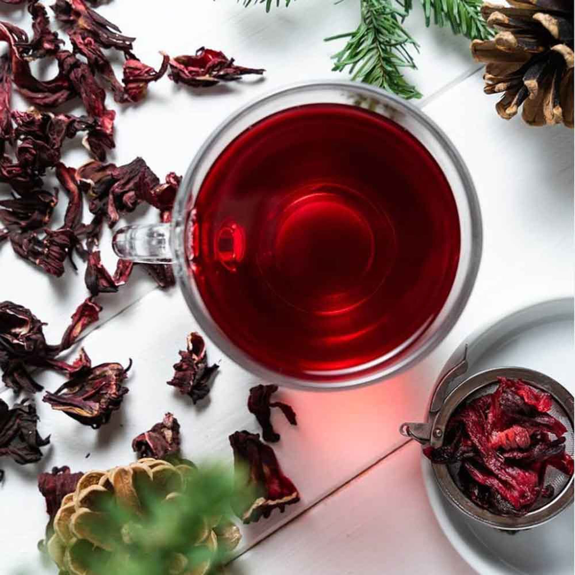 Una tazza di tè all'ibisco rosso, con fiori secchi e un colino da tè accanto.
