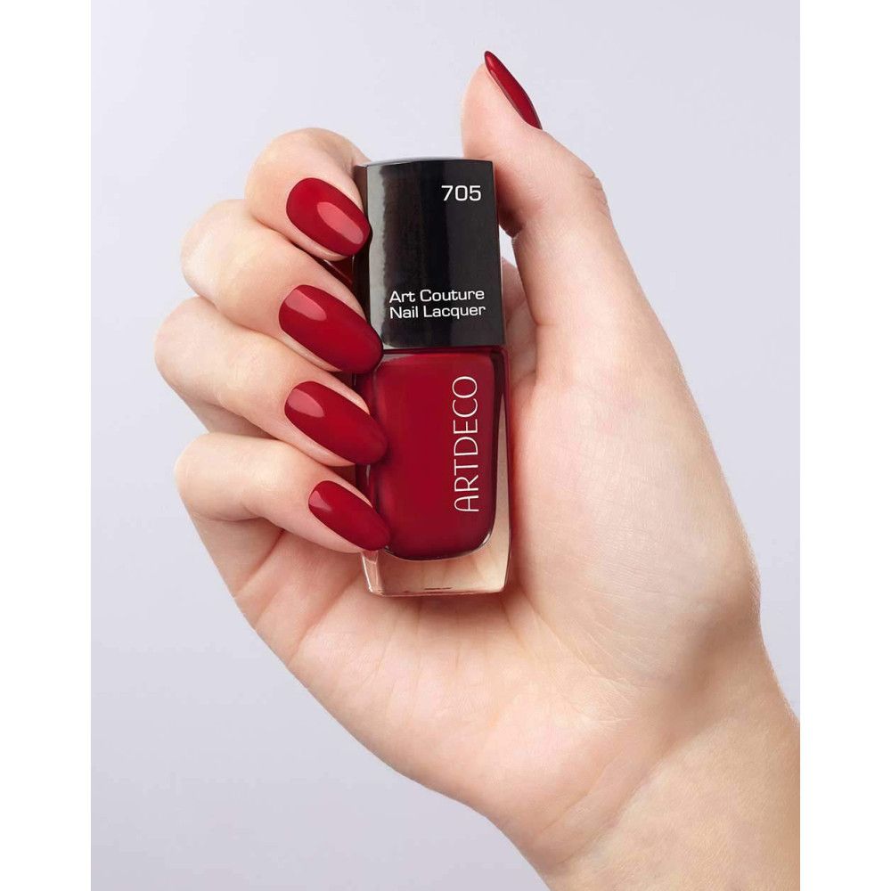 Mano con un flacone di smalto rosso. Flacone con tappo nero, scritta ARTDECO e Art Couture Nail Lacquer. Unghie smaltate.