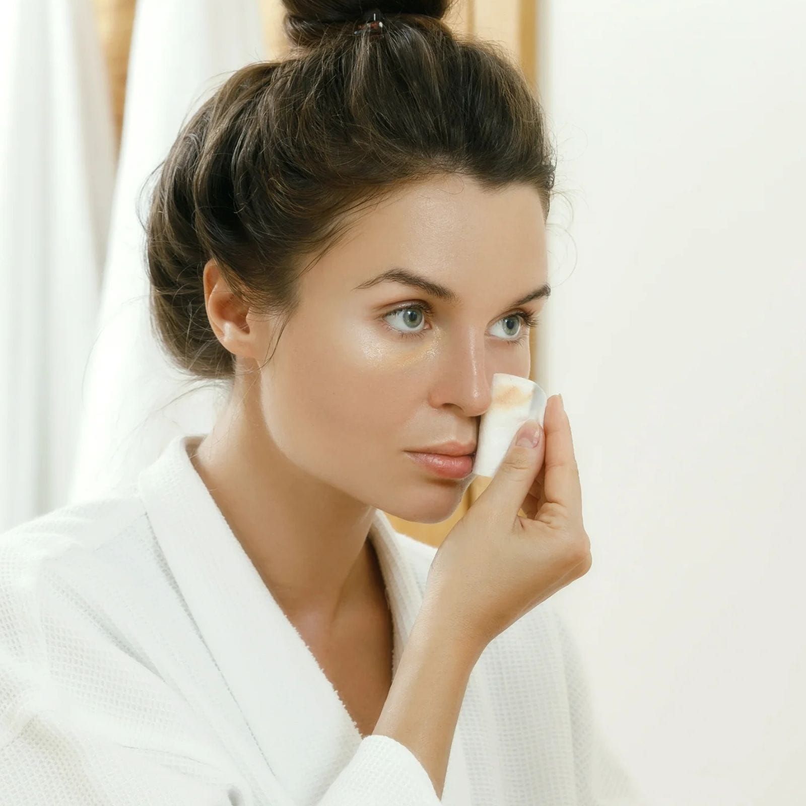 Una donna con indosso un accappatoio bianco applica il fondotinta sul viso usando una spugnetta, concentrandosi sulla zona sotto gli occhi.