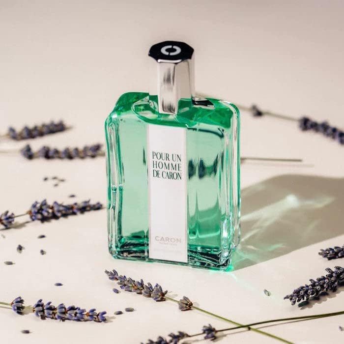 Flacone di vetro verde con tappo argentato. Scritta: Pour Un Homme de Caron. Circondato da fiori di lavanda su sfondo chiaro.