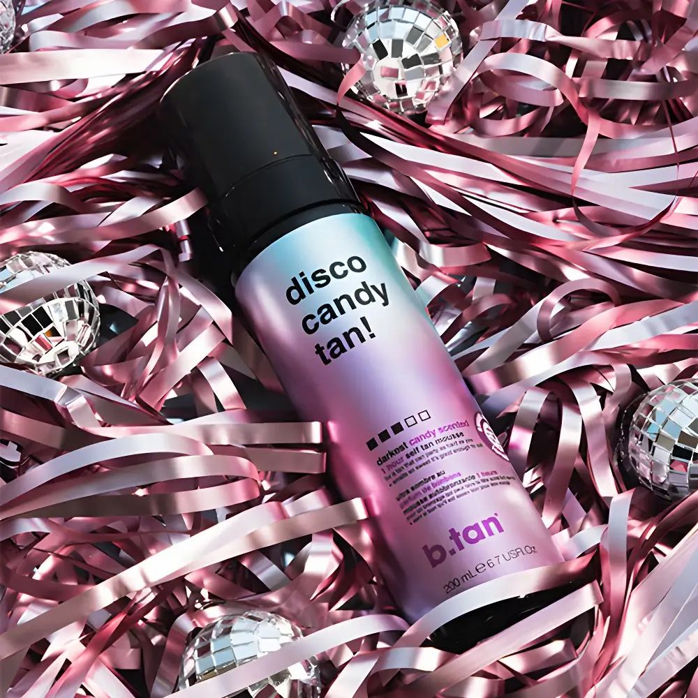 Flacone di mousse autoabbronzante su coriandoli rosa e sfere a specchio. Erogatore nero, etichetta sfumata con nome e loghi.