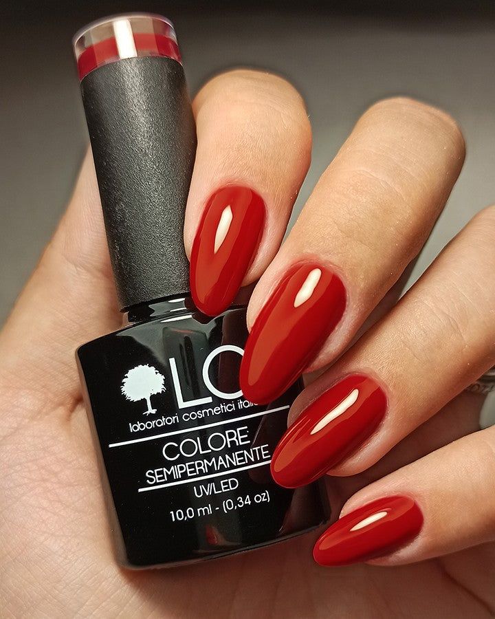 Mano con unghie smaltate e flacone di smalto. Unghie rosse e flacone di smalto nero. Scritta: LCI, Colore Semipermanente.