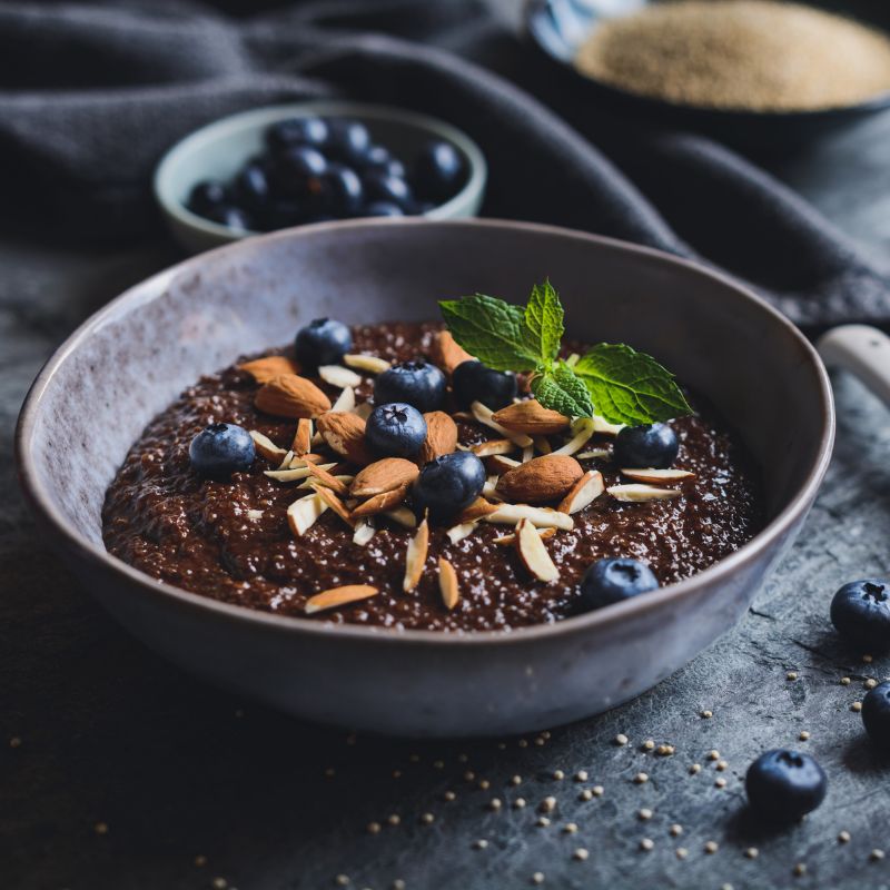 Ciotola con porridge marrone, guarnito con mirtilli, mandorle e menta. Su sfondo scuro.
