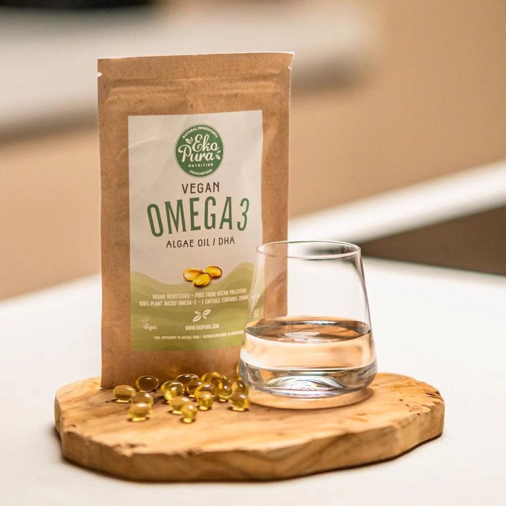 Confezione marrone con stampa. Accanto, un bicchiere d'acqua e capsule su tagliere in legno. Scritta: Vegan Omega 3 Algae Oil.
