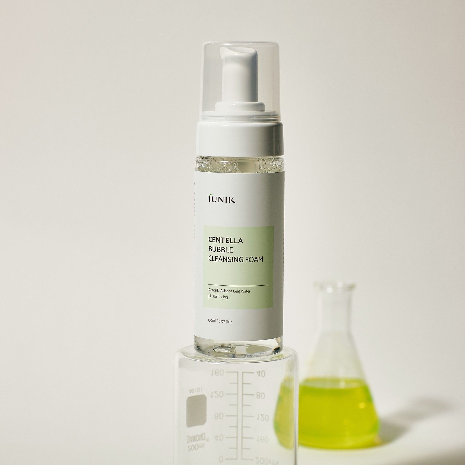Flacone iUNIK Centella Bubble Cleansing Foam su un cilindro graduato. Un becher con liquido giallo è accanto.