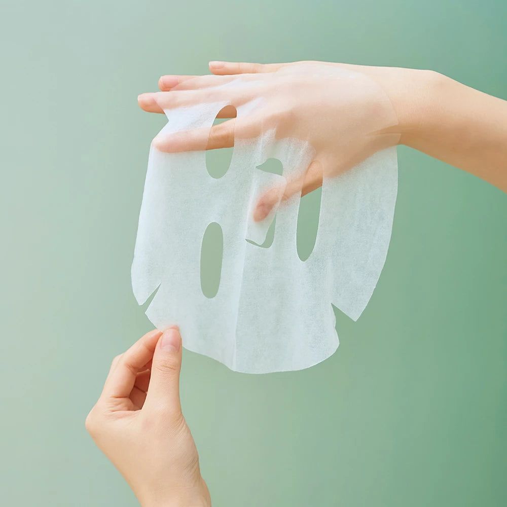 Mani che tengono una maschera per il viso bianca. La maschera è trasparente e di forma ovale. Lo sfondo è verde.