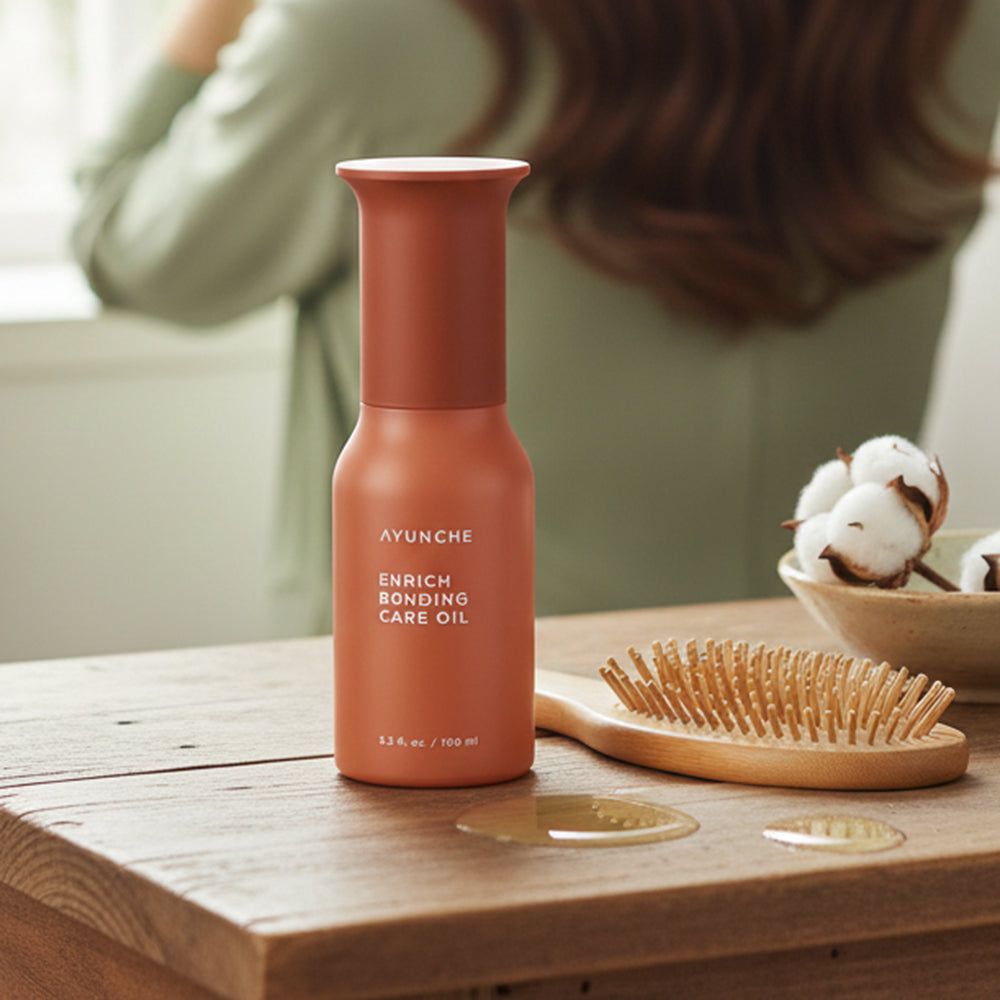 Flacone AYUNCHE Enrich Bonding Care Oil accanto a una spazzola per capelli e batuffoli di cotone su un tavolo di legno.