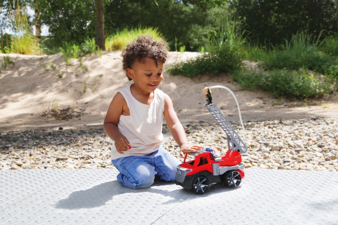 Un bambino gioca con un'autopompa giocattolo rossa all'aperto. Il bambino è seduto per terra e sorride.