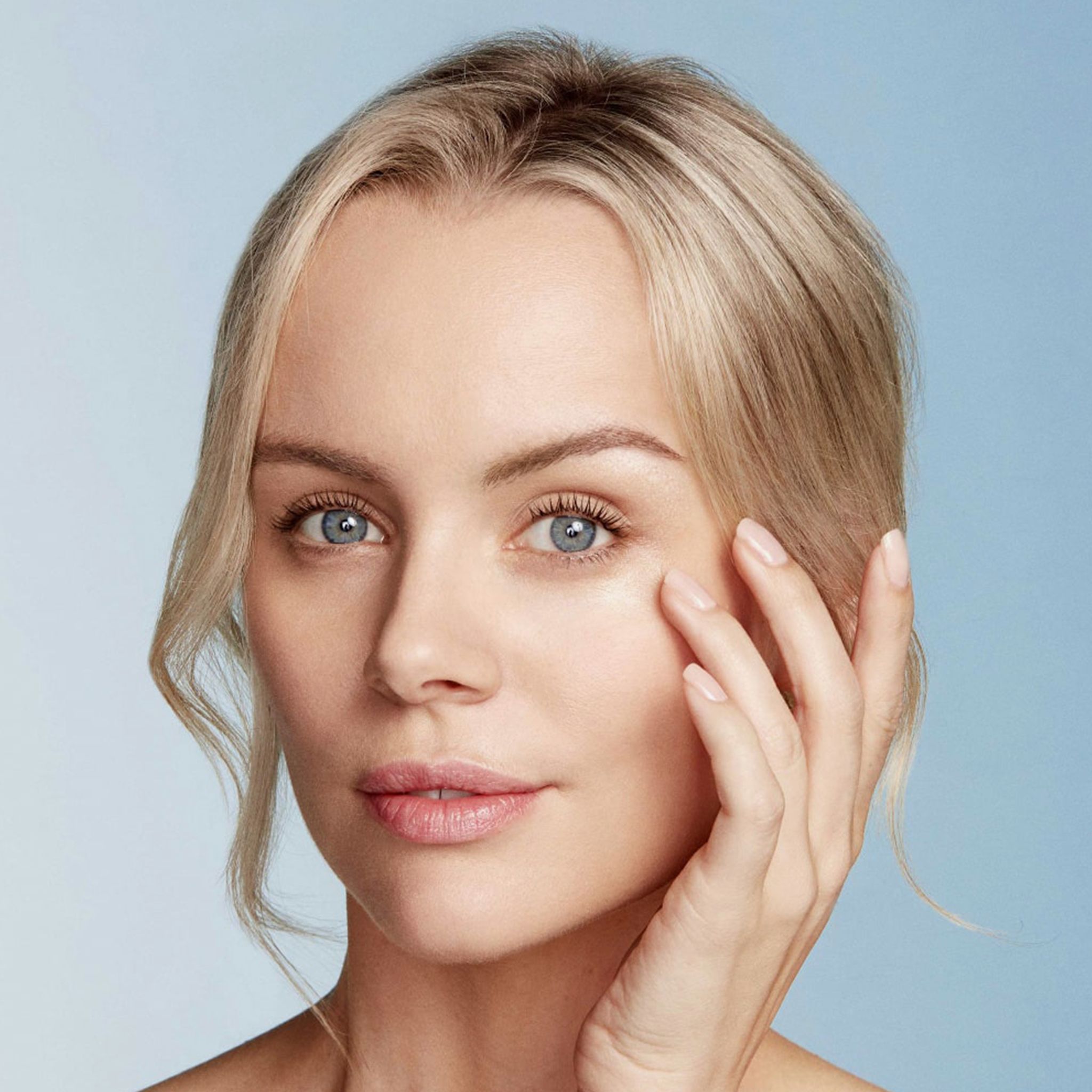 Donna bionda tocca delicatamente la pelle sotto l'occhio. Sfondo blu. Nessuna indicazione del prodotto visibile.