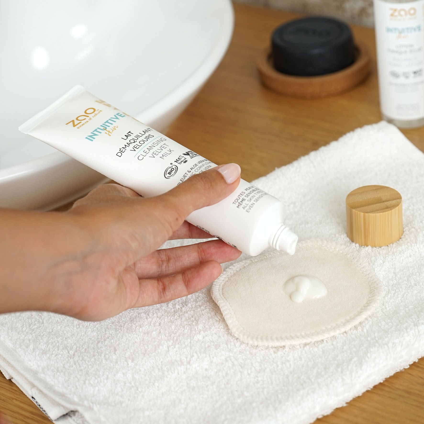 Tubo bianco con prodotto, testo e logo. Prodotto versato su un dischetto. Su asciugamano e vicino al lavabo.