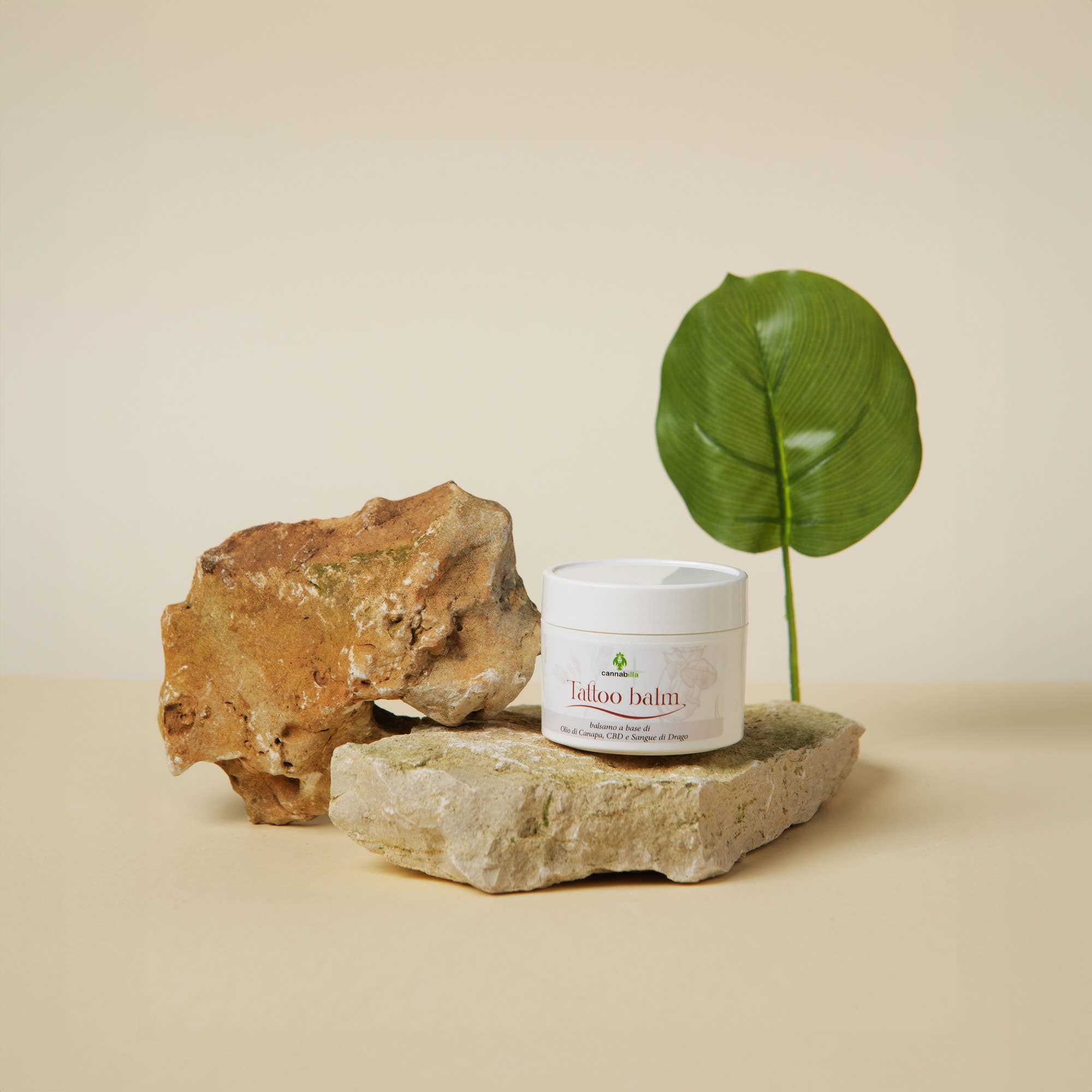 Vasetto bianco con scritta "Tattoo balm" su pietre e foglia. Scritta "balsamo a base di Olio di Canapa, CBD e Sangue di Drago".