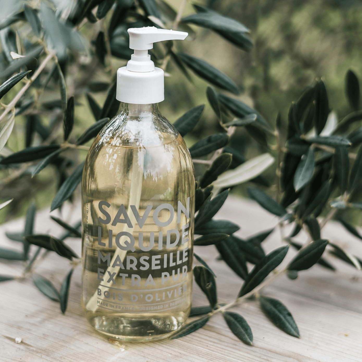 Flacone trasparente con dosatore davanti a rami di ulivo. Scritta: Savon Liquide Marseille Extra Pur Bois d'Olivier.