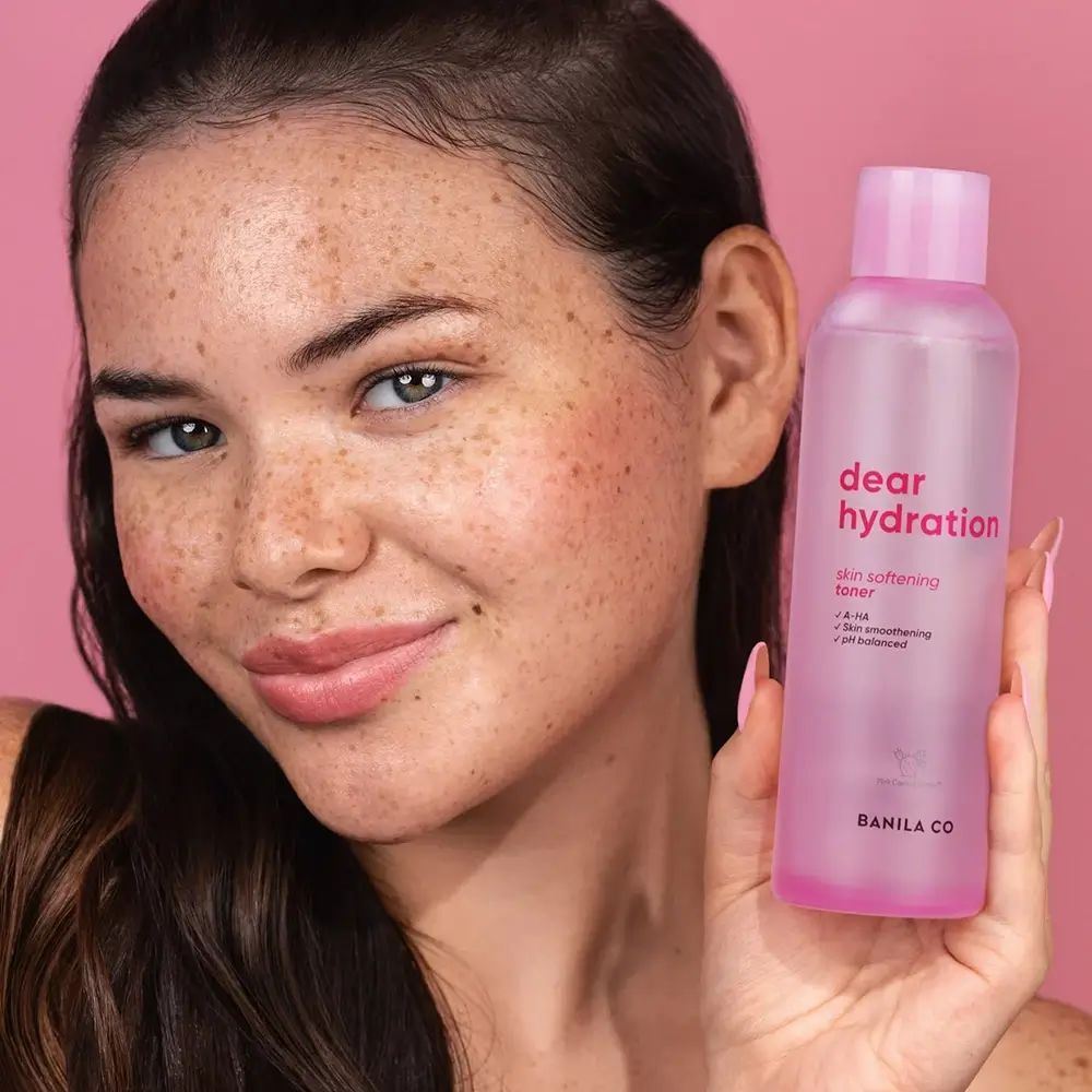 Flacone rosa con scritta "dear hydration" e "skin softening toner". Donna che tiene il flacone su sfondo rosa.