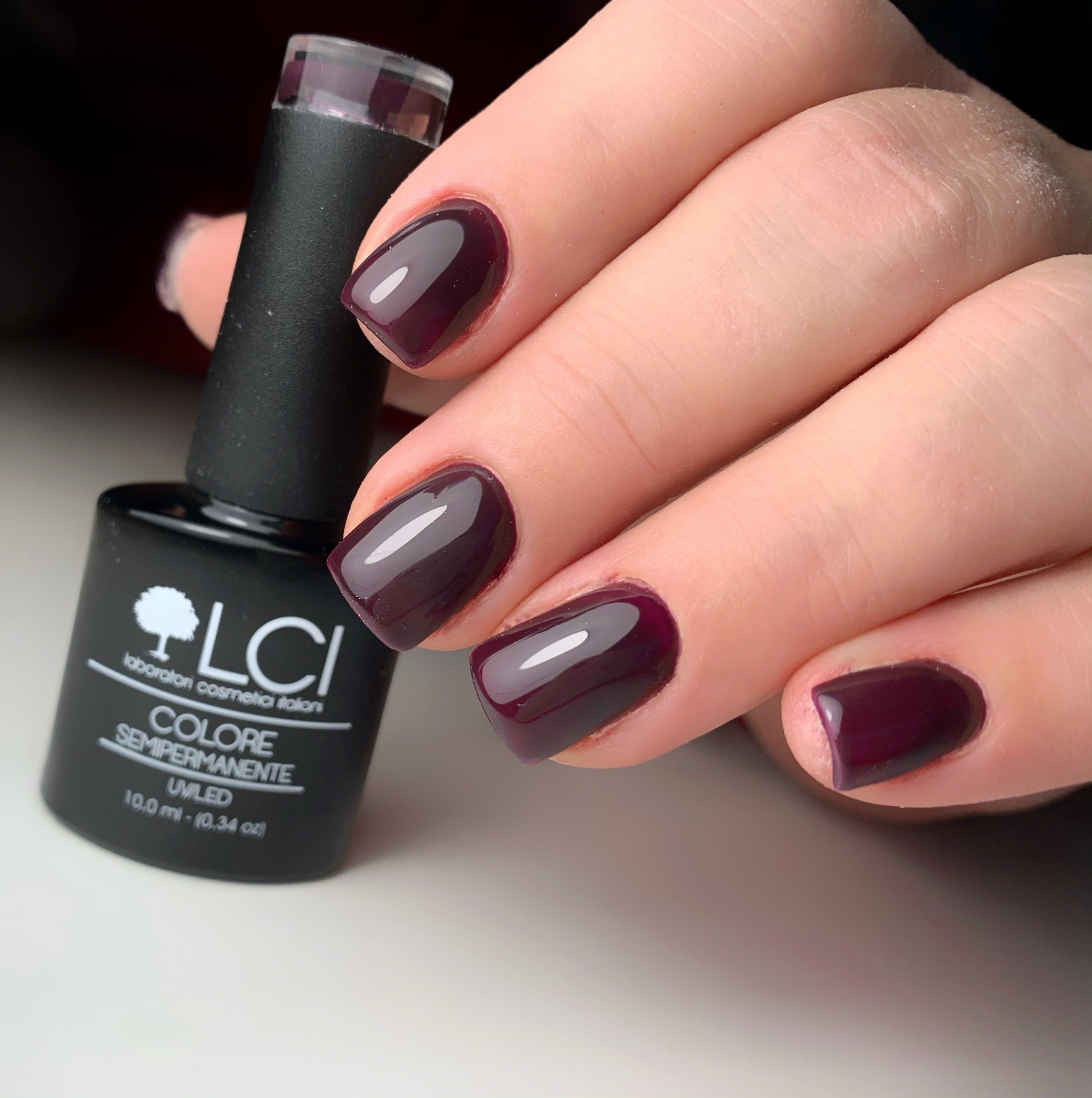 Mano con unghie smaltate e flacone di smalto. Colore: viola scuro. Scritte: LCI, COLORE SEMIPERMANENTE, HEMA FREE.