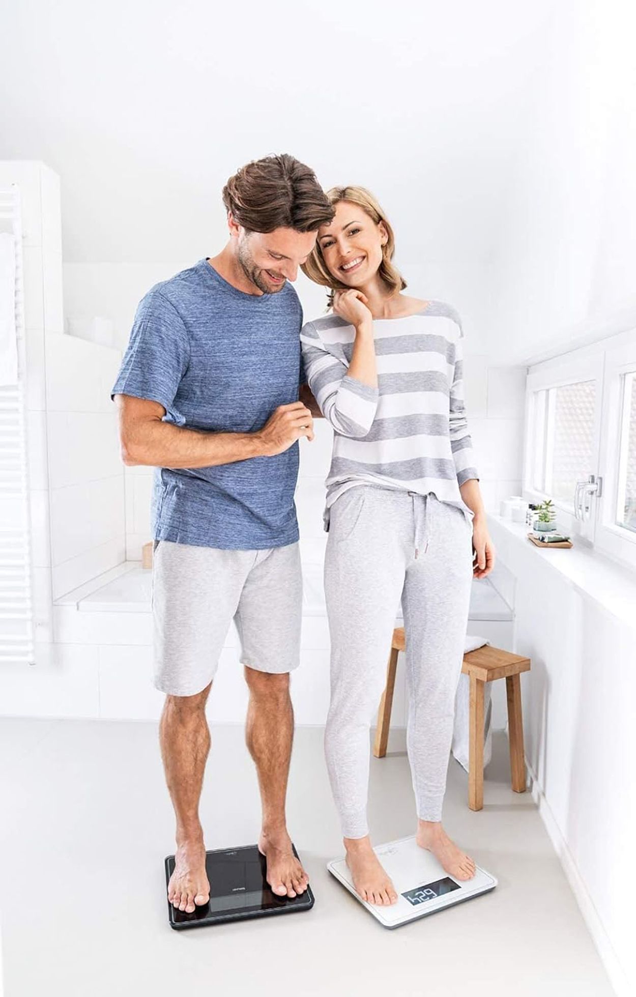 Una coppia in piedi su bilance. Entrambe mostrano un peso. Sfondo bagno. Bilance nere e bianche.