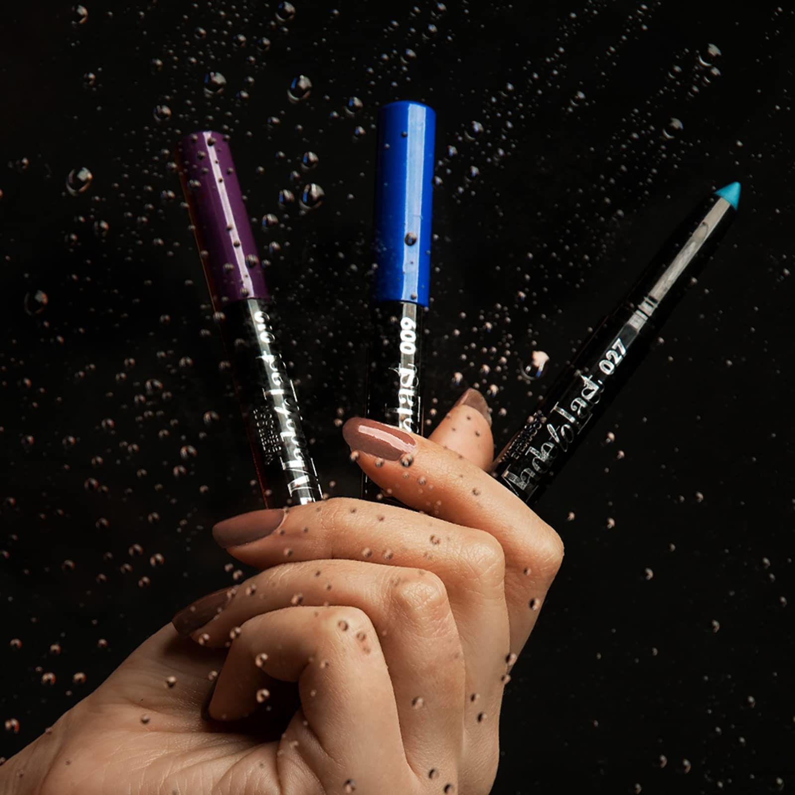 Tre matite ombretto di diversi colori. Le matite sono tenute da una mano. Gocce d'acqua sullo sfondo.