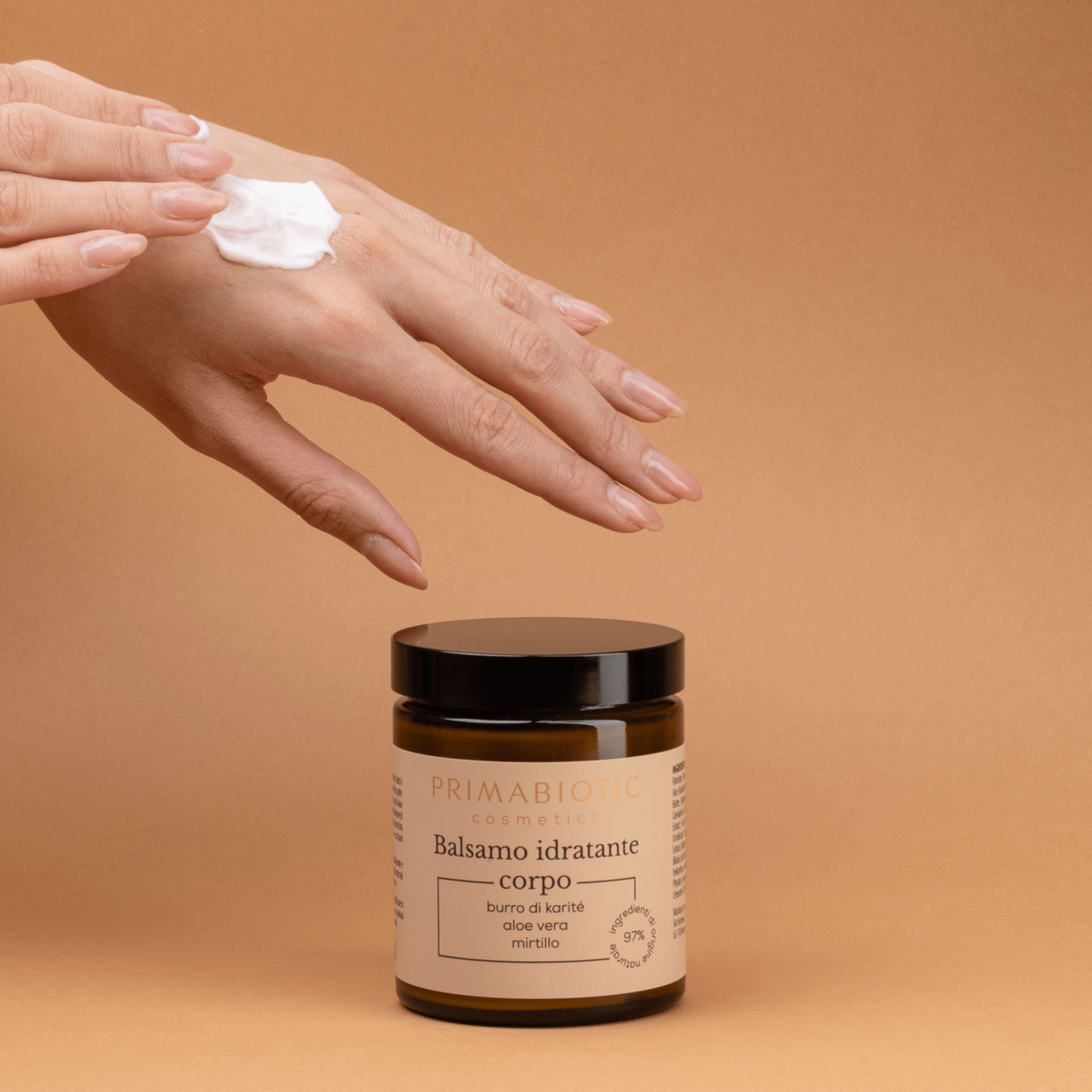 Mano con crema sulla pelle. Vaso in vetro marrone con coperchio nero. L'etichetta recita: Balsamo idratante corpo, Primabiotic Cosmetics.
