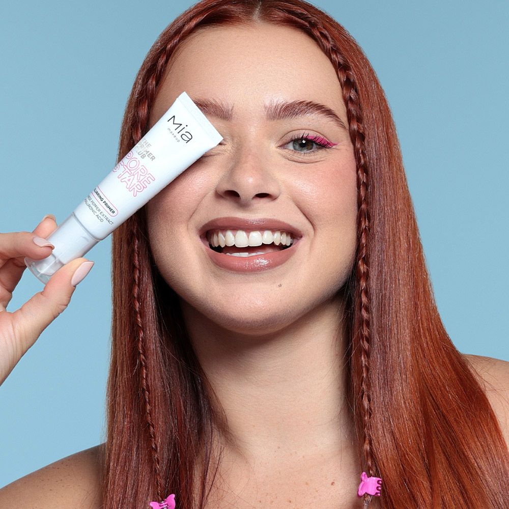Donna tiene in mano un tubetto di Mia Makeup Pore Star Primer. Tubo bianco con scritte rosa. Persona con trecce rosse e eyeliner rosa.