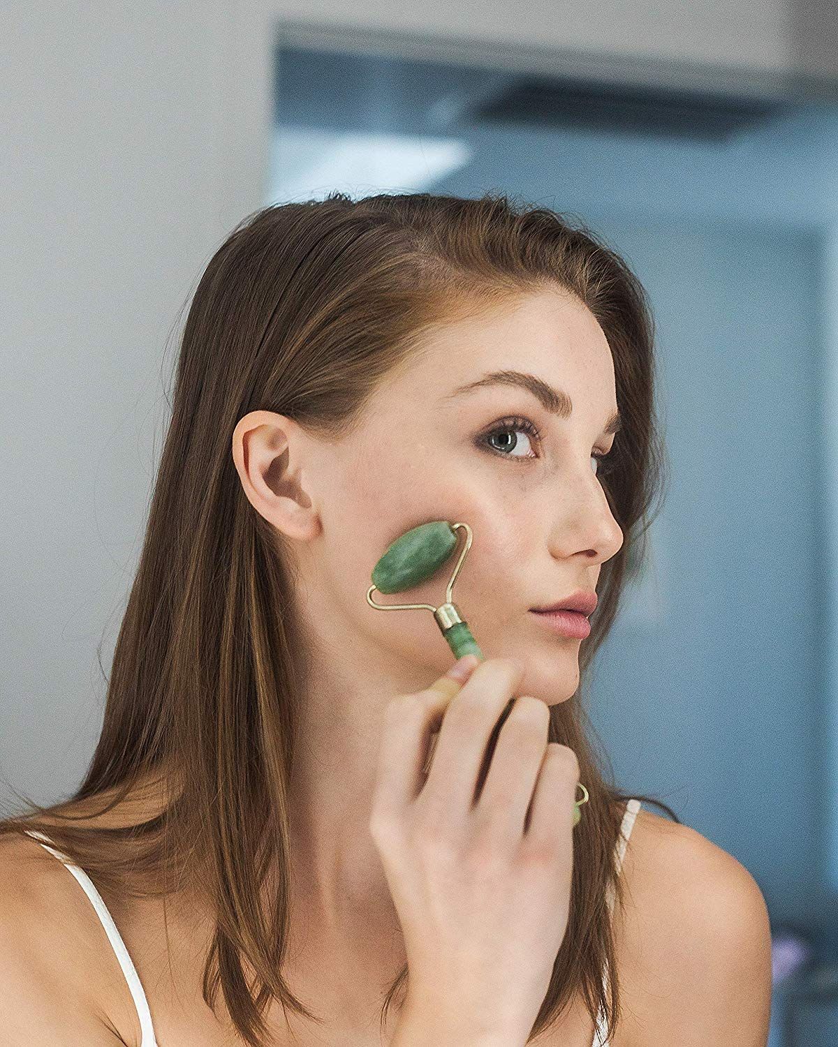 Una donna con i capelli castani usa un rullo facciale di giada verde sulla guancia.