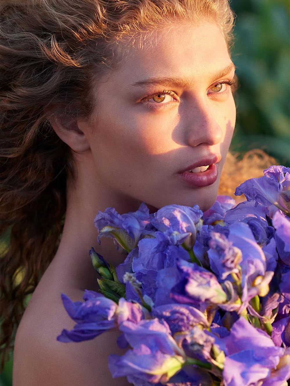 Ritratto di donna con ricci che tiene un mazzo di iris viola. Luce naturale, primo piano.