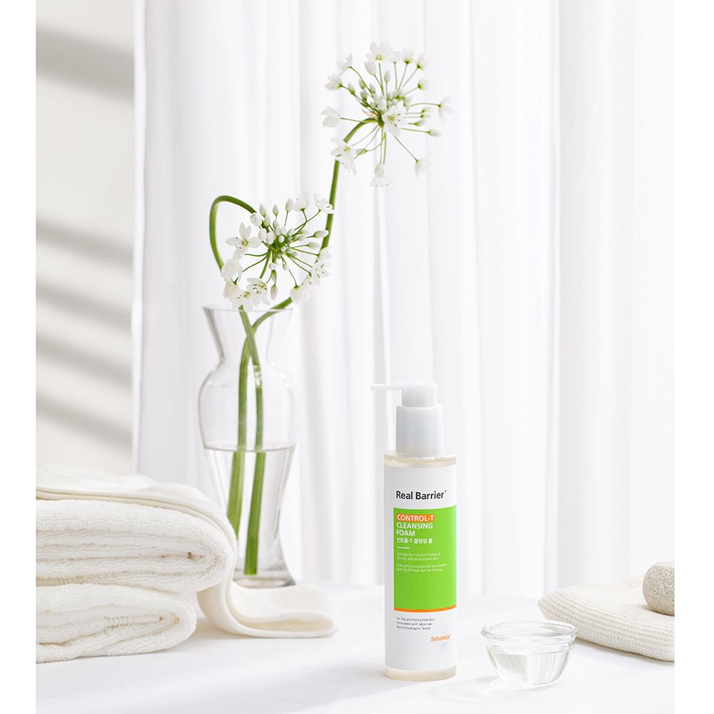 Flacone bianco-verde con dosatore vicino a fiori e asciugamani. Testo: Real Barrier, Control-T Cleansing Foam. Ciotola con liquido.