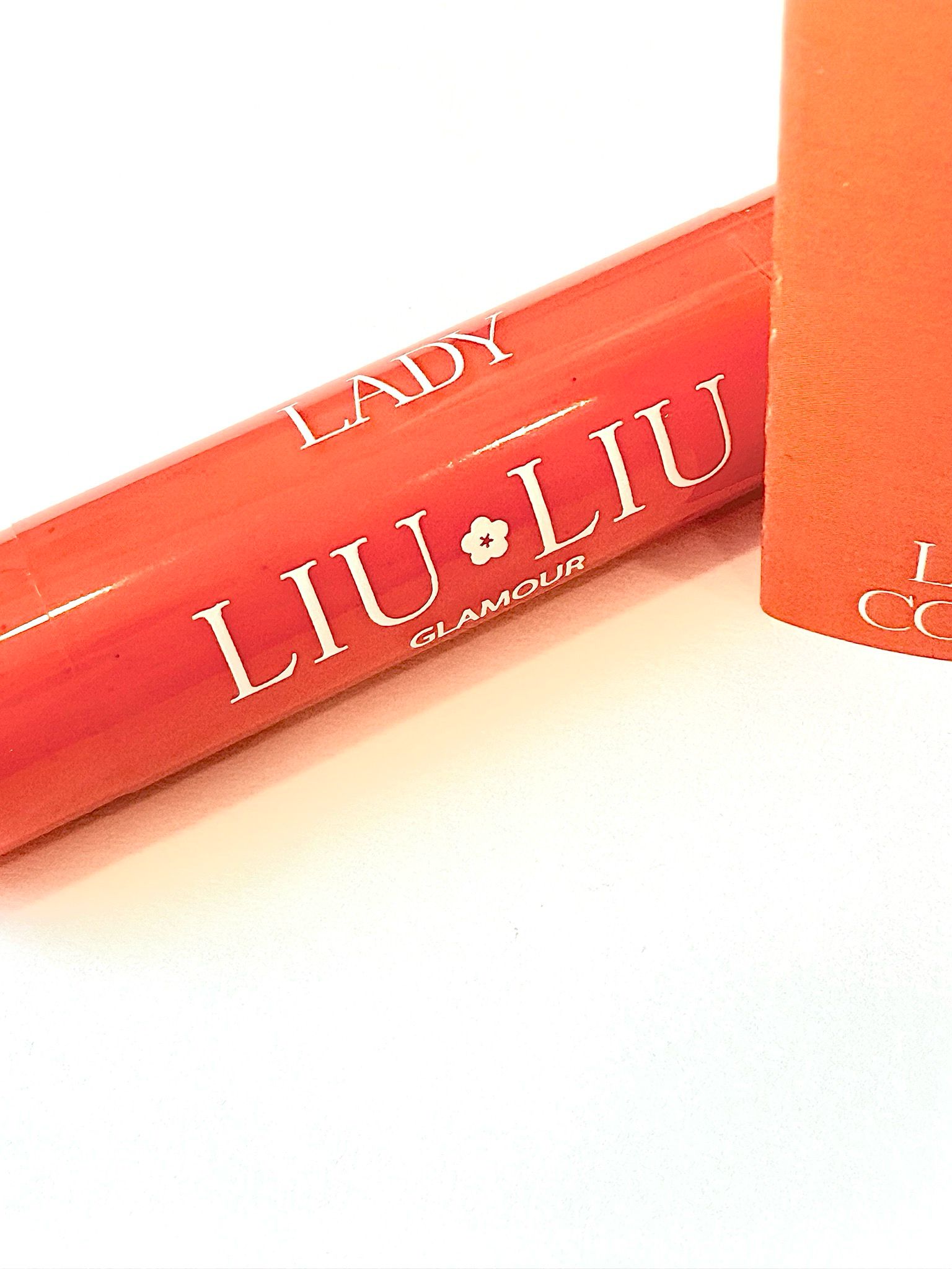 Primo piano di un tubetto di lucidalabbra. Scritta: LADY LIU LIU GLAMOUR. Sfondo arancione.