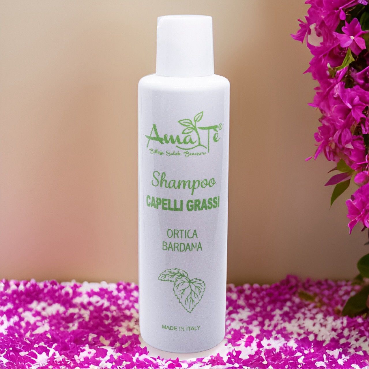 Flacone bianco con etichetta verde. Testo: Amatè Shampoo Capelli Grassi Ortica Bardana. Sfondo: petali.