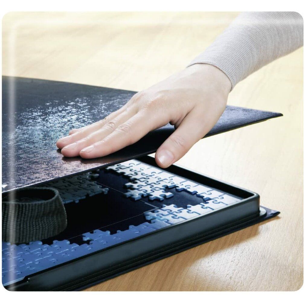 Mano che posiziona un tappetino per puzzle su un contenitore nero. Pezzi parzialmente visibili. Manico nero.