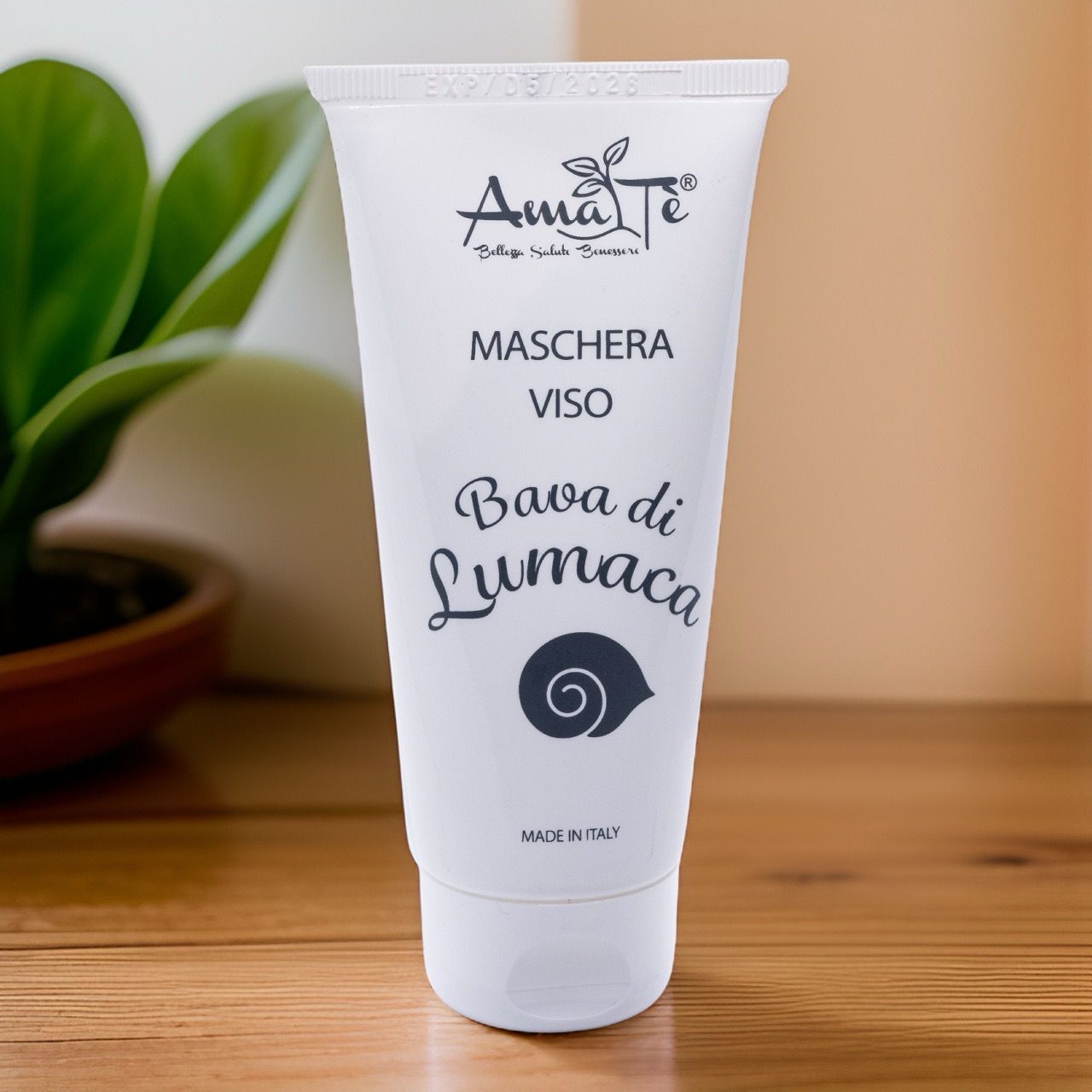 Tubo bianco con logo Amatè e testo: Maschera Viso Bava di Lumaca. Sfondo: legno e pianta.