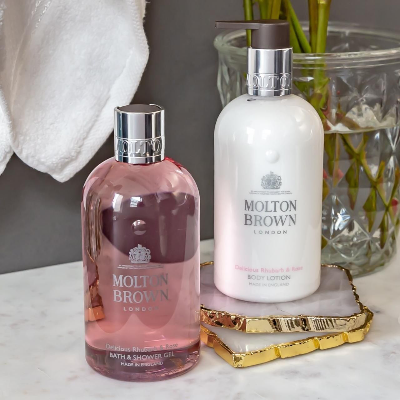 Due prodotti Molton Brown su un vassoio. Gel bagno e doccia e lozione per il corpo. Sfondo: asciugamano, vaso con fiori.