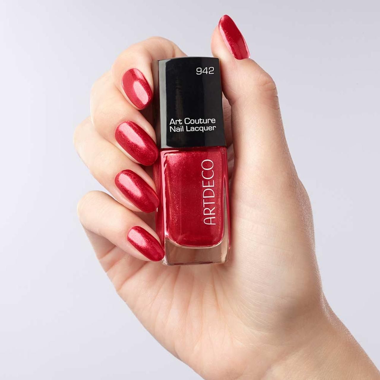Mano che tiene un flacone di smalto e unghie smaltate. Smalto rosso, scritta: Art Couture Nail Lacquer, ARTDECO, 942.