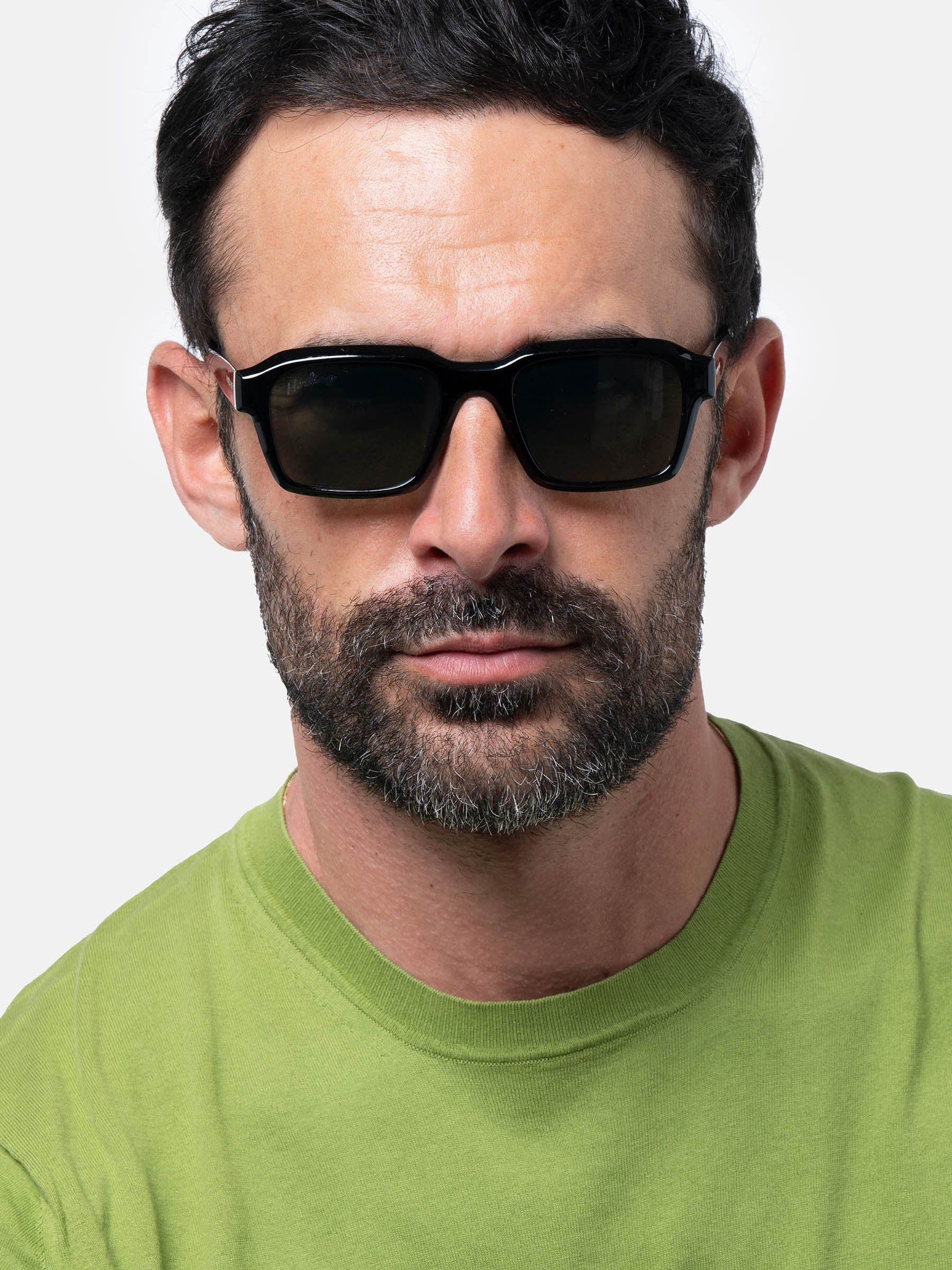 Uomo con occhiali da sole neri. Forma rettangolare, lenti verdi. Barba e maglietta verde.