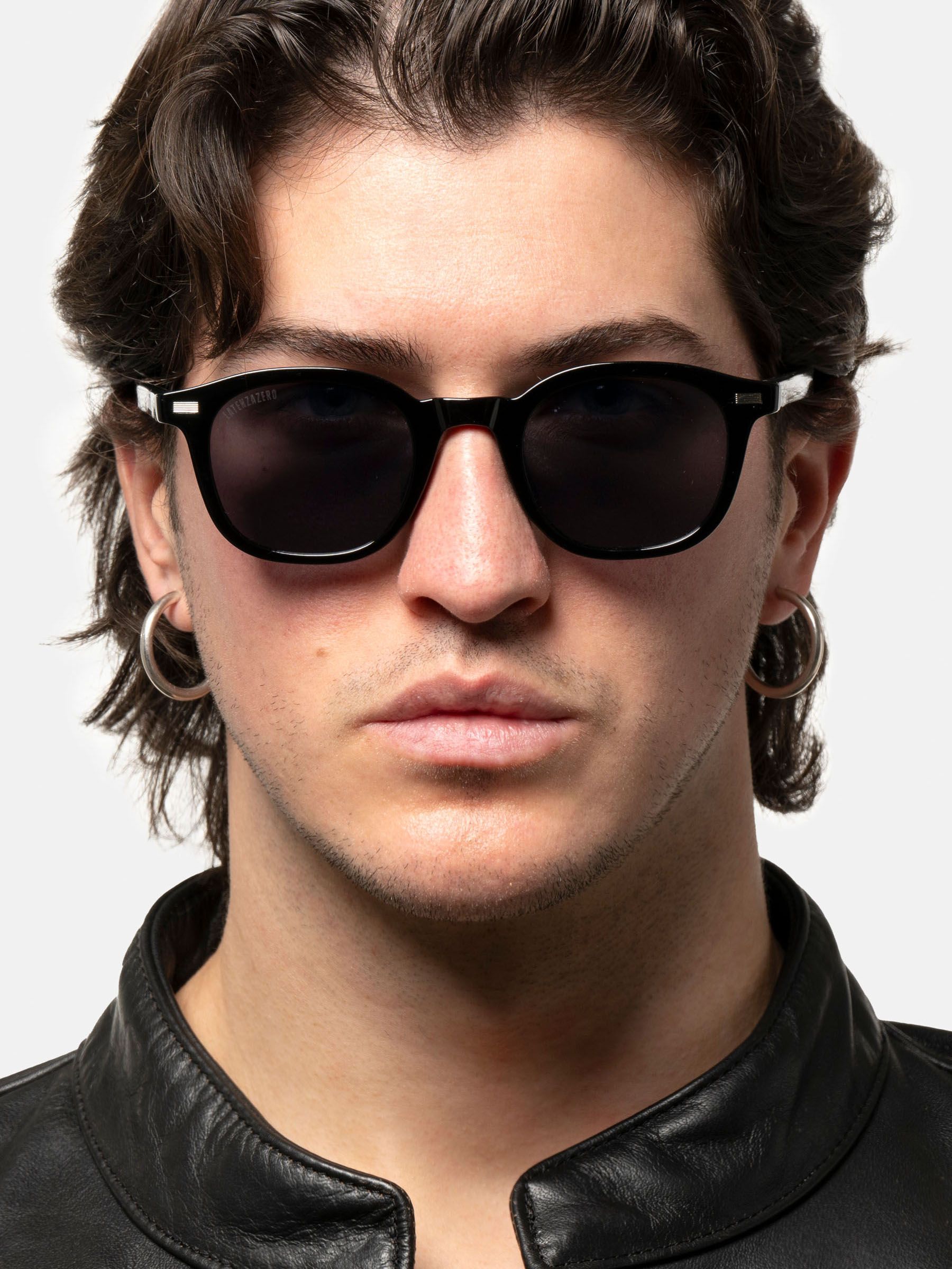 Uomo con occhiali da sole neri. Lenti rotonde, montatura nera. Orecchini d'argento. Marchio sull'asta.