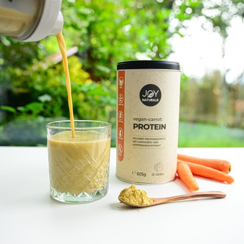 Bicchiere con bevanda, accanto barattolo, carote e polvere su cucchiaio. Scritta: JOY NATURALS, vegan-carrot:PROTEIN, 605g. Complesso proteico vegano.