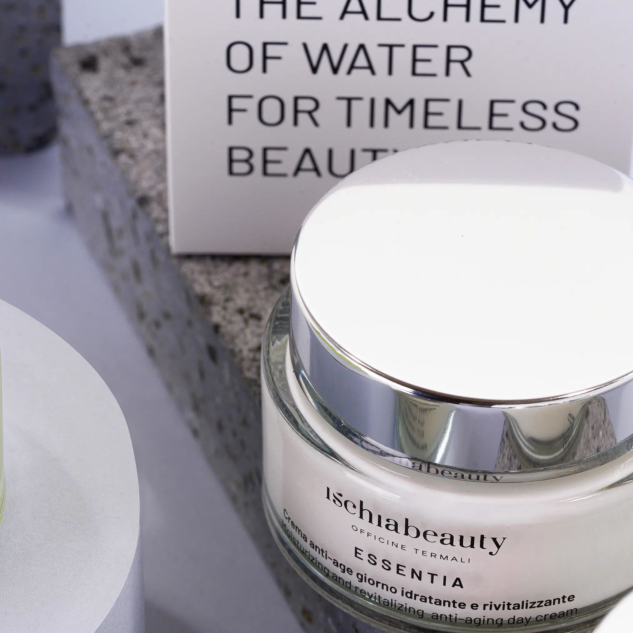 Vaso di crema con coperchio argentato. Scritta: ischiabeauty, ESSENTIA, Crema anti-age giorno. Testo sulla confezione sullo sfondo.