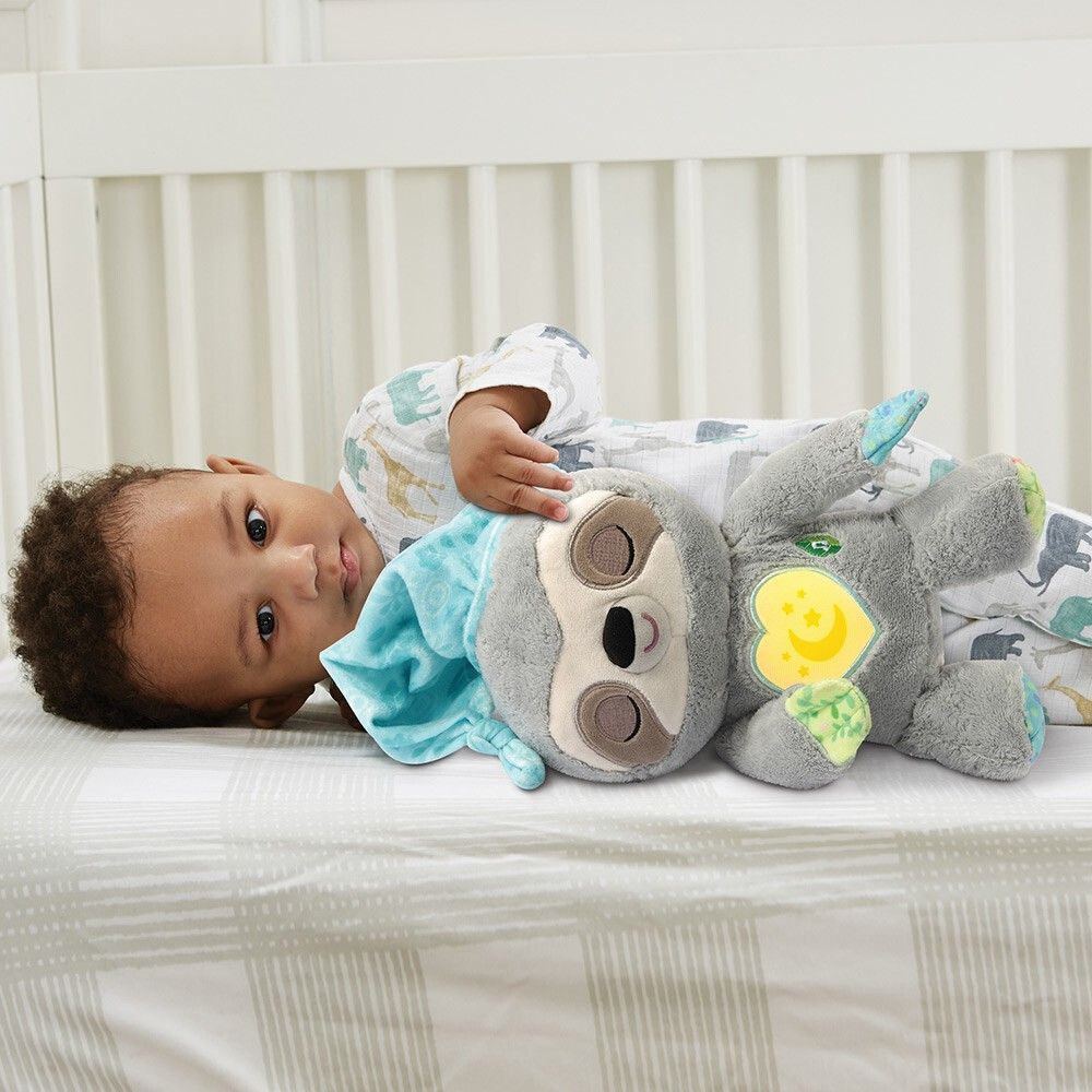 Bambino sdraiato sulla schiena nel letto accanto a un peluche bradipo grigio con cappello. Cuore giallo con motivo luna-stella.