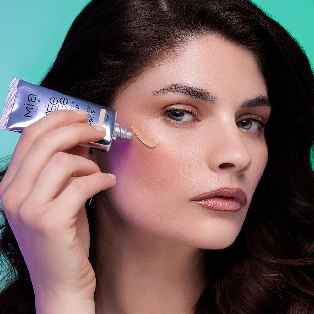 Una donna applica un fondotinta chiaro SPF 15 da un tubetto argentato sulla guancia.