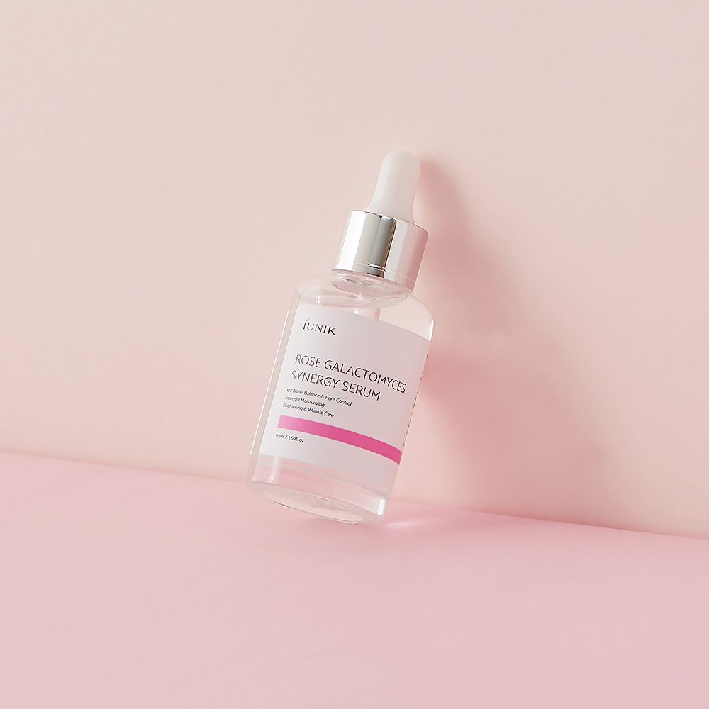 Flacone di vetro trasparente con pipetta. Scritta: ROSE GALACTOMYCES SYNERGY SERUM. Striscia rosa e testo. Su sfondo rosa.